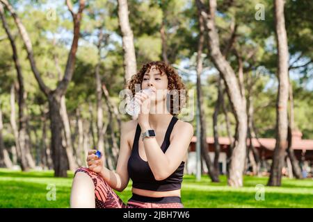 Bella donna rossa che indossa abiti sportivi in piedi sul parco cittadino, acqua potabile all'aperto per rinfrescarsi con il sorriso sul viso. Sport all'aperto, guarire Foto Stock