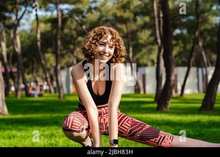 Donna sportiva e carina che indossa abiti sportivi nel parco cittadino, all'aperto allunga i muscoli delle gambe, facendo affondi laterali in posizione skandasana. Stile di vita sano co Foto Stock