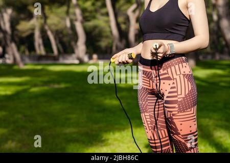 Bella donna rossa che indossa un reggiseno sportivo nero in piedi sul parco cittadino, all'aperto a stomaco piatto con una corda che salta nelle sue mani a livello della vita. Salute Foto Stock