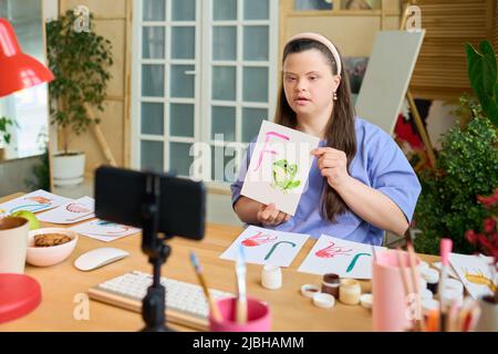 Giovane donna con sindrome di Down che mostra carta con rana verde dipinta e lettera F mentre comunica con il pubblico online Foto Stock