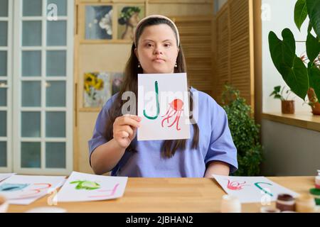 Giovane insegnante serio che mostra carta con lettera verde disegnata J e medusa rossa mentre si siede dalla scrivania di fronte al computer o alla macchina fotografica Foto Stock