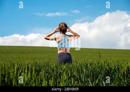 Vista posteriore di una donna sportiva e in forma in piedi in un campo verde indossando un paio di cuffie per ascoltare musica. Foto Stock