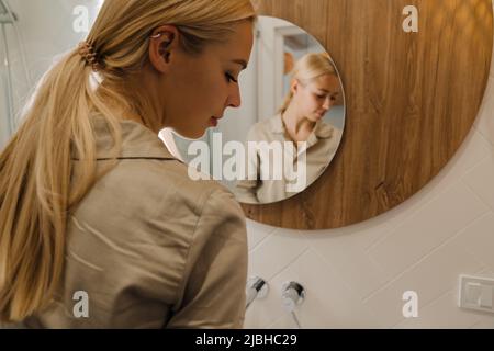 Giovane donna bionda che guarda nello specchio mentre si lava in bagno Foto Stock