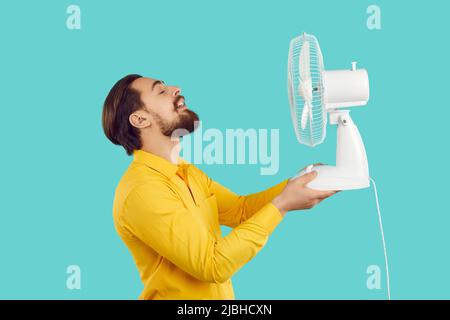 L'uomo bearded divertente gode l'aria fresca e scappa il calore con la ventola elettrica su sfondo azzurro chiaro. Foto Stock