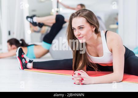 Cute sorridente giovane donna in sportswear facendo avanzato esercizio di divisione centrale che si piega sopra appoggiandosi sulle braccia in palestra. Foto Stock