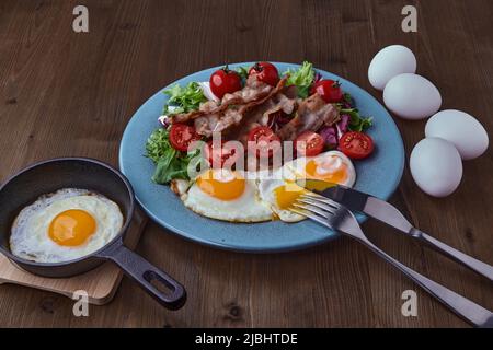 Uova fritte con pancetta, lattuga e pomodori ciliegini su un piatto blu su un tavolo scuro. Padella con uova fritte e uova intere di pollo. Giorno delle uova Foto Stock