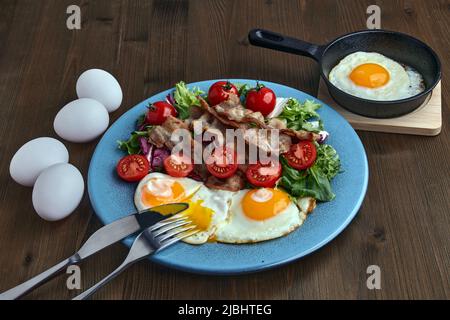 Uova fritte con pancetta, lattuga e pomodori ciliegini su un piatto blu su un tavolo scuro. Padella con uova fritte e uova intere di pollo. Foto Stock