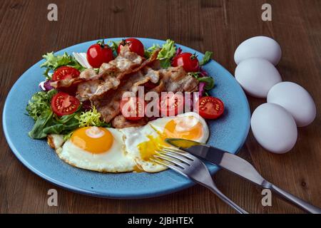 Uova fritte con pancetta, lattuga e pomodori ciliegini su un piatto blu con posate e uova intere di pollo. Giorno delle uova strapazzate Foto Stock