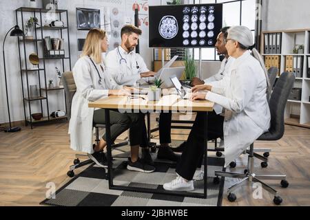 Gruppo di quattro lavoratori medici pensivi che guardano la prescrizione del paziente e che considerano i modi di trattamento della salute. Colleghi multiculturali seduti attorno al tavolo con display che mostra l'immagine TC cerebrale. Foto Stock