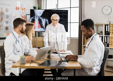 Medici senior esperti ponting sul monitor con scansione a raggi X e frequenza heartbeat durante la conferenza con i colleghi. Gruppo di operatori medici che hanno il brainstorming nella moderna sala riunioni. Foto Stock