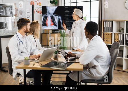 Lavoratore medico anziano che discute con colleghi multirazziali scansione a raggi X e risultati heartbeat mostrati sul grande schermo digitale. Team di medici che hanno briefing in sala di consiglio di amministrazione circa paziente severo. Foto Stock