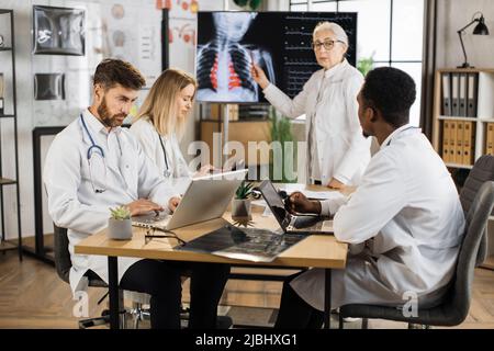 Dottore qualificato che conduce seminario a tre interns multirazziale nella sala riunioni. Donna anziana in piedi vicino allo schermo digitale e spiegare i risultati della scansione radiologica toracica. Foto Stock