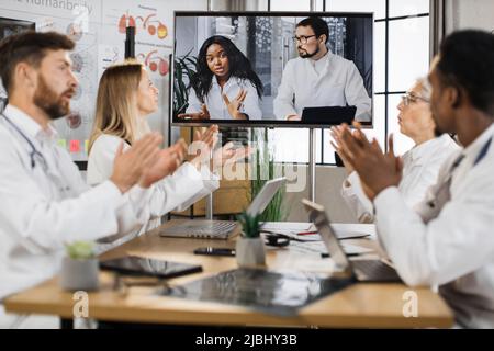 Gruppo di medici maschi e femmine che allietano con applausi i loro colleghi multiculturali durante la presentazione medica online in clinica. Attenzione allo schermo digitale moderno. Foto Stock