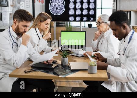 Gruppo di quattro operatori medici pensivi che guardano la presunzione del paziente e che considerano i modi di soluzione del problema. Colleghi multiculturali che utilizzano un computer portatile con schermo a tasti cromati mentre lavorano in ospedale. Foto Stock