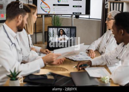 Gruppo di personale di medicina concentrato in uniformi bianche di laboratorio seduti al tavolo della clinica durante la lezione online di ingegneria genetica. Focus su scienziata afro-americana su schermo laptop. Foto Stock