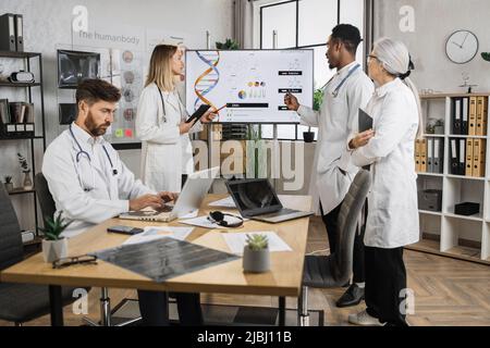 Gruppo di medici multietnici concentrati in piedi vicino allo schermo durante la sessione di lavoro nella sala riunioni del laboratorio. Maschio caucasico che lavora su un laptop mentre il team di colleghi che parla di ingegneria genetica. Foto Stock