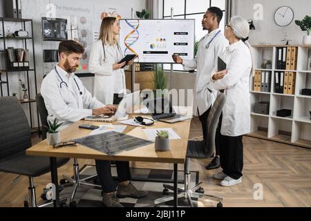 Gruppo di medici multietnici concentrati in piedi vicino allo schermo durante la sessione di lavoro nella sala riunioni del laboratorio. Maschio caucasico che lavora su un laptop mentre il team di colleghi che parla di ingegneria genetica. Foto Stock