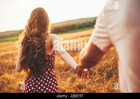 Vista posteriore di una donna zenzero irriconoscibile che tiene la mano del suo ragazzo e guida l'uomo attraverso il campo di grano. Tempo libero, viaggi, turismo. Foto Stock