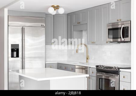 Una cucina grigia con elettrodomestici in acciaio inossidabile, rubinetto d'oro, ripiani in marmo bianco e un backsplash di piastrelle della metropolitana. Foto Stock