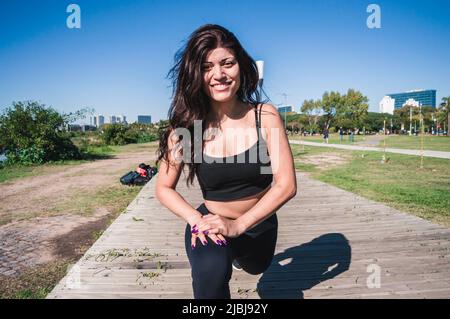 donna latina caucasica sportiva all'aperto, gambe allunganti che riscaldano i muscoli, guardando la macchina fotografica e sorridendo, cielo dietro di lei. Foto Stock