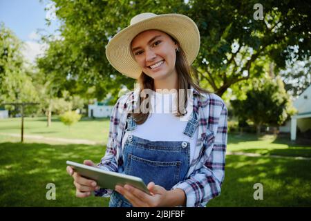 Donna contadina caucasica che indossa un cappello di paglia in piedi all'aperto con tablet digitale Foto Stock