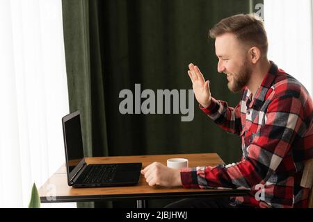 Sorridente bell'uomo che ha una videochiamata in bar, uomo d'affari bearded che parla alla videochiamata con i colleghi, freelance che prende l'ordine dal cliente via Foto Stock