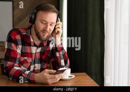 Ritratto di bel bearded uomo cuffie guardare video telefono cellulare moderno studio loft. L'uomo è seduto su una sedia, tenendo uno smartphone e. Foto Stock