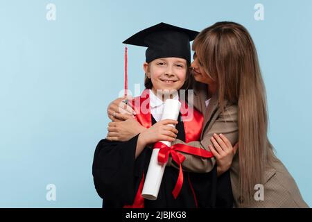 Piccola ragazza laureata per celebrare la laurea. Bambino con cappuccio di laurea e certificato di tenuta dell'accappatoio per cerimonie. Mamma abbraccia e congratulazioni figlia Foto Stock