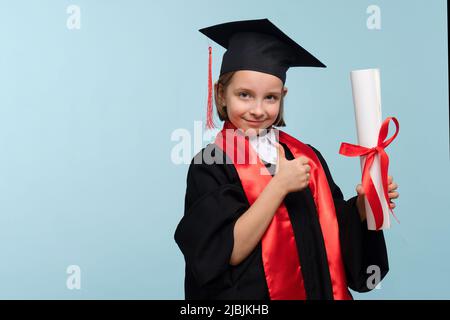 Bambina di 9-11 anni con cappuccio di laurea e accappatoio per cerimonie con diploma certificato. La bambina laureata ha emozioni positive, mostrando il pollice Foto Stock