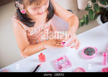 Asian adorabile divertente ragazza che fa makeup tuffi pennello in bottiglia per dipingere unghie lucidare vernice rossa chiodo lei stessa, attività di apprendimento per essere donna, Foto Stock