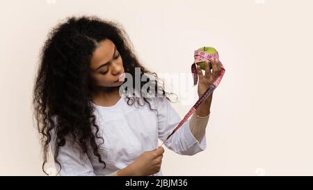 African American Female Nutrizionista guarda mela verde e nastro di misurazione in mani. Foto Stock
