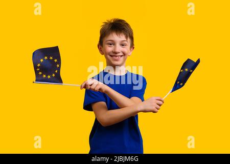 Happy schoolboy in Blue Basic T-shirt che ondeggiava due bandiere dell'Unione europea su sfondo giallo in Studio Foto Stock