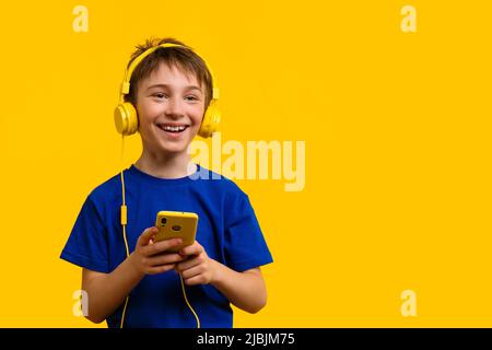 Ragazzo positivo 9-11y.o in T-shirt blu di base e cuffie gialle con smartphone. I sorrisi dello schoolboy mantengono ampiamente le mani sul telefono delle cellule che è in movimento Foto Stock