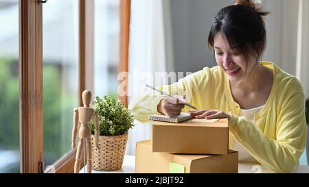 Indirizzo di scrittura femminile sorridente su scatola di cartone e preparazione per la spedizione. E-commerce, concetto di vendita online Foto Stock