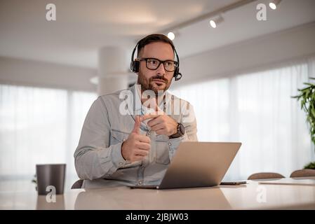 Uomo d'affari serio che indossa cuffie e gesturing. Il dirigente maschio che indossa casuals astuti sta lavorando sul laptop. È seduto alla scrivania in ufficio. Foto Stock