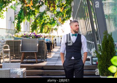 Giovane uomo dall'aspetto hipster in un abito con una cravatta ad arco in una caffetteria estiva Foto Stock