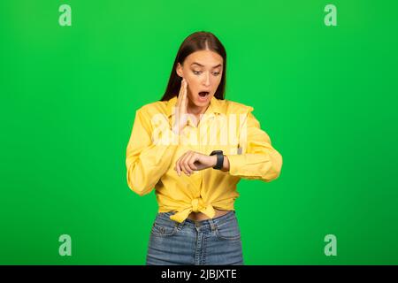 Shocked Millennial Lady guardando Smartwatch su Green Studio sfondo Foto Stock