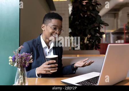 La donna felice seduta in un caffè con una bevanda calda fa una riunione in linea, videochiamata, guarda la macchina fotografica del laptop e sorride, condivide le buone notizie al lun Foto Stock
