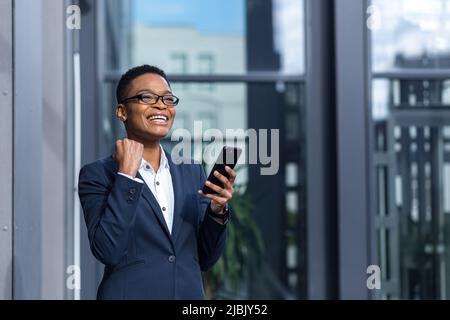 Felice e riuscita donna di affari gioisce nel giorno di vittoria e felice, celebra, sorridente africano americano tiene il telefono nelle mani Foto Stock