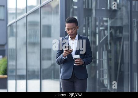 Una signora fiduciosa afroamericana cammina intorno al quartiere degli affari e utilizza il telefono. Tenere una tazza di caffè, vestita con abiti formali. Attrattiva Foto Stock