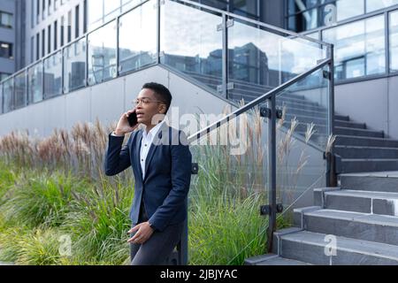 Donna d'affari arrabbiata e seria che parla al telefono fuori dell'ufficio, donna afroamericana vestita in vestiti d'affari, comunicante con la collega Foto Stock