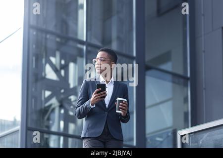 Una signora fiduciosa afroamericana cammina intorno al quartiere degli affari e utilizza il telefono. Tenere una tazza di caffè, vestita con abiti formali. Attrattiva Foto Stock