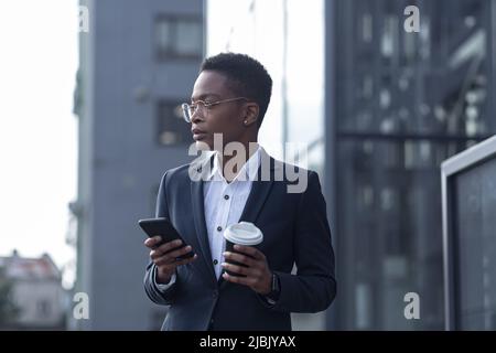 Una signora fiduciosa afroamericana cammina intorno al quartiere degli affari e utilizza il telefono. Tenere una tazza di caffè, vestita con abiti formali. Attrattiva Foto Stock