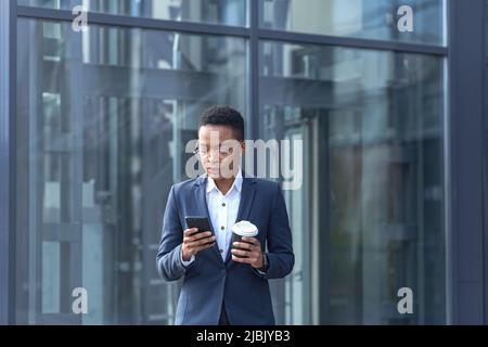 Una signora fiduciosa afroamericana cammina intorno al quartiere degli affari e utilizza il telefono. Tenere una tazza di caffè, vestita con abiti formali. Attrattiva Foto Stock