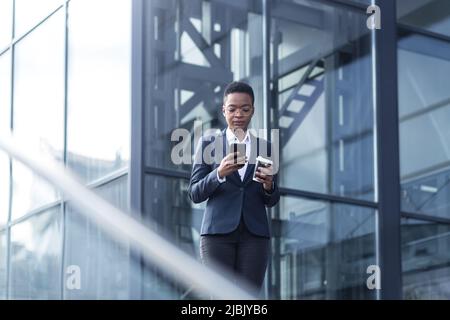 Una signora fiduciosa afroamericana cammina intorno al quartiere degli affari e utilizza il telefono. Tenere una tazza di caffè, vestita con abiti formali. Attrattiva Foto Stock