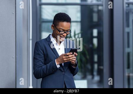 Felice e riuscita donna di affari gioisce nel giorno di vittoria e felice, celebra, sorridente africano americano tiene il telefono nelle mani Foto Stock