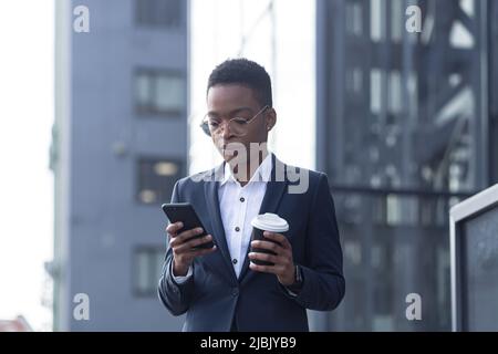 Una signora fiduciosa afroamericana cammina intorno al quartiere degli affari e utilizza il telefono. Tenere una tazza di caffè, vestita con abiti formali. Attrattiva Foto Stock