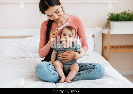 La madre che si prende cura di spazzolare i capelli della figlia, il bambino seduto sui giri della mamma e guardando la macchina fotografica, la donna che ama l'infanzia Foto Stock