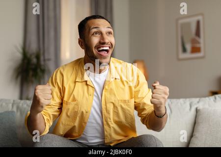 Il giovane uomo nero eccitato gesturing sì, celebrando il successo o la grande vittoria, seduto sul divano a casa Foto Stock