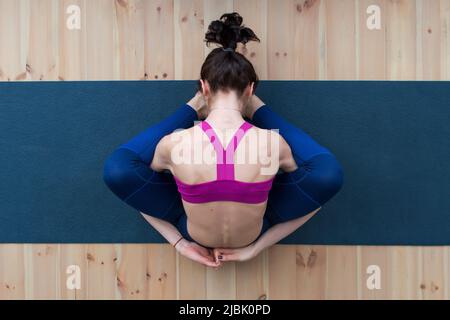 Vista dall'alto della giovane donna caucasica seduta in dormiente tartaruga posa suppta kurmasana sul pavimento in studio. Foto Stock